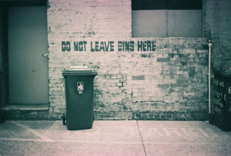 A bleak image of a No Parking space, with a sign that says 'Do not leave bins here'. There is a bin directly under the sign.