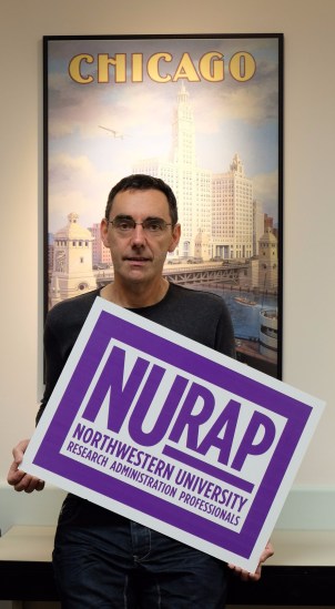 Jonathan looking nervous, as he holds a NURAP sign in front of a poster that says 'Chicago'