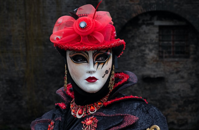 Person wearing a gorgeous white-faced mask with elaborate eye make-up and deep red lips, along with a fancy red hat and elaborate clothing and jewels. 