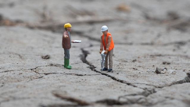 Two tiny figures working on a crack in the footpath. One has a jackhammer and a high-viz vest. The other has a clipboard. 